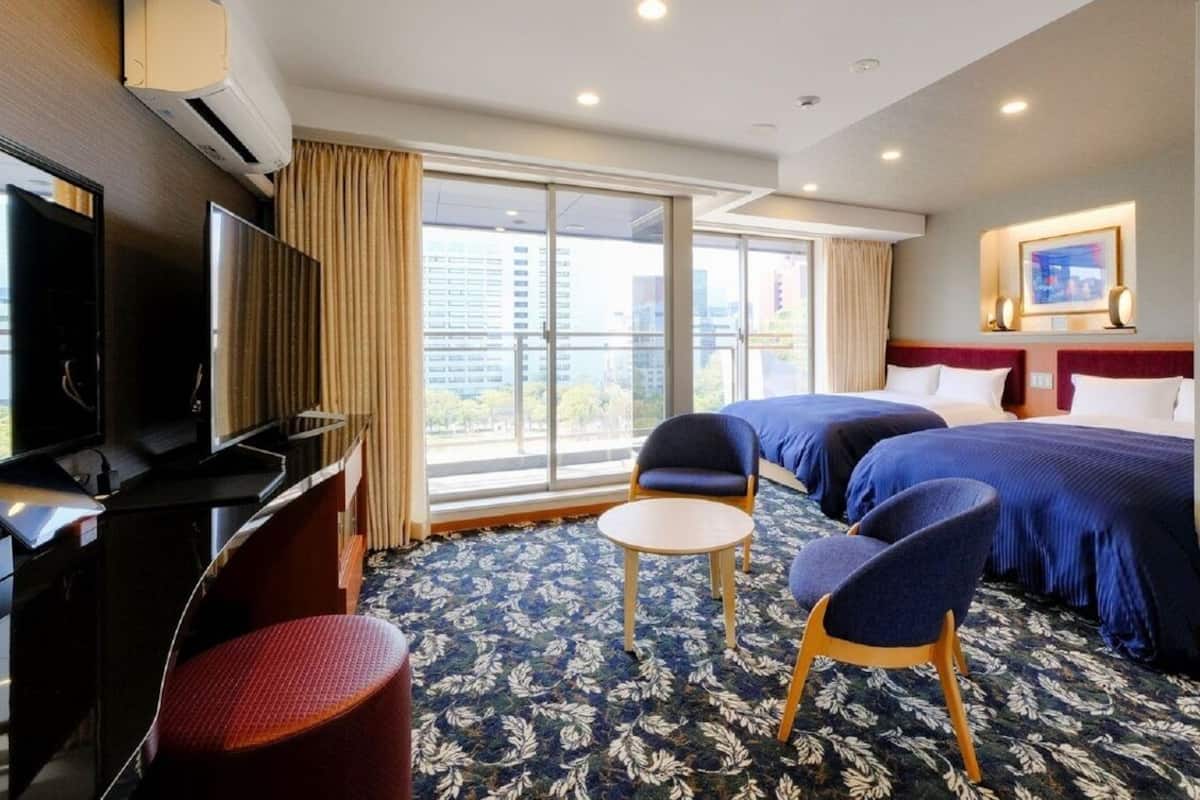 Habitación de hotel con dos camas, televisión, una mesita, una silla y un ventanal con vistas a la ciudad.