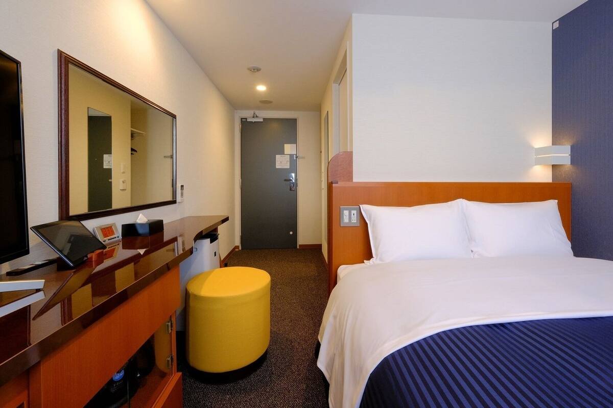 Ein Hotelzimmer mit Bett, Schreibtisch mit Computer, Spiegel, Mülleimer und Tür.
