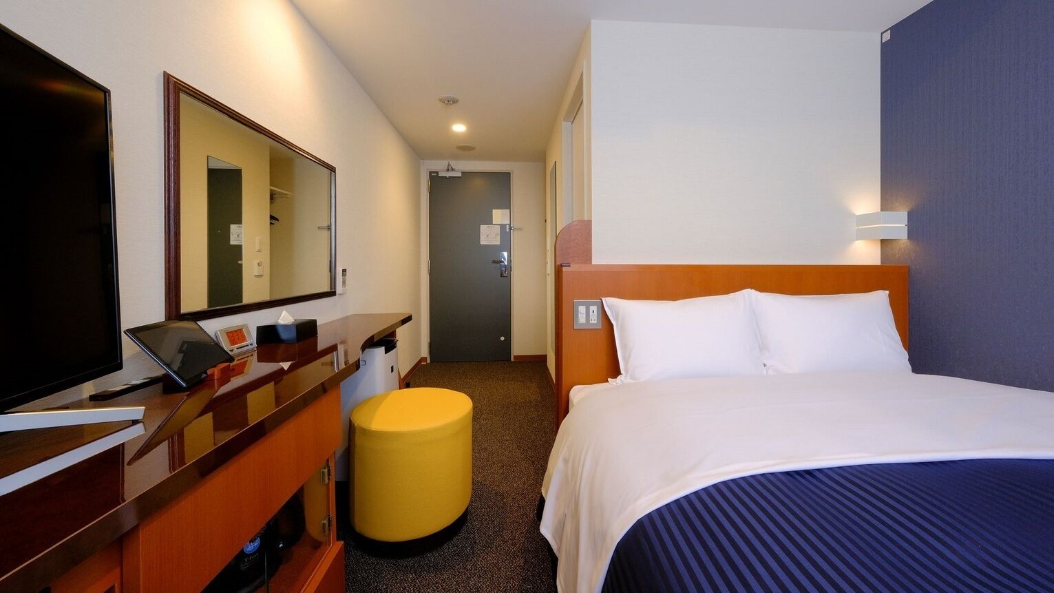Una habitación de hotel con una cama, un escritorio con una computadora, un espejo y un taburete pequeño.