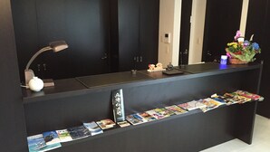 Un comptoir d’accueil avec un plan de travail noir, une lampe blanche, une petite statue, un vase avec des fleurs et une rangée de magazines.