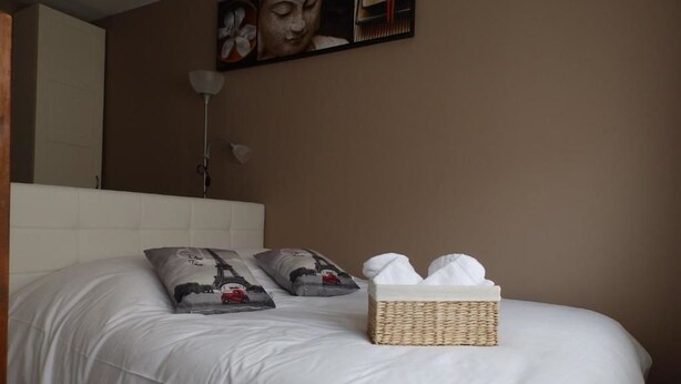 A neatly made bed with white bedding, a decorative pillow, a small wicker basket with towels, and a wall-mounted lamp. - Lens Louvre 2 (Lens)