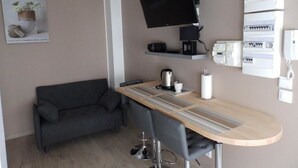 A compact living space with a sofa, a small dining table, a TV mounted on the wall, and a kitchenette area with electrical outlets and a coffee machine. - Lens Louvre 2 (Lens)