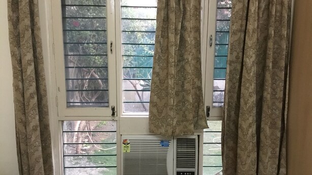 Et vindue med mønstrede gardiner, en synlig aircondition og grøn vegetation udenfor.