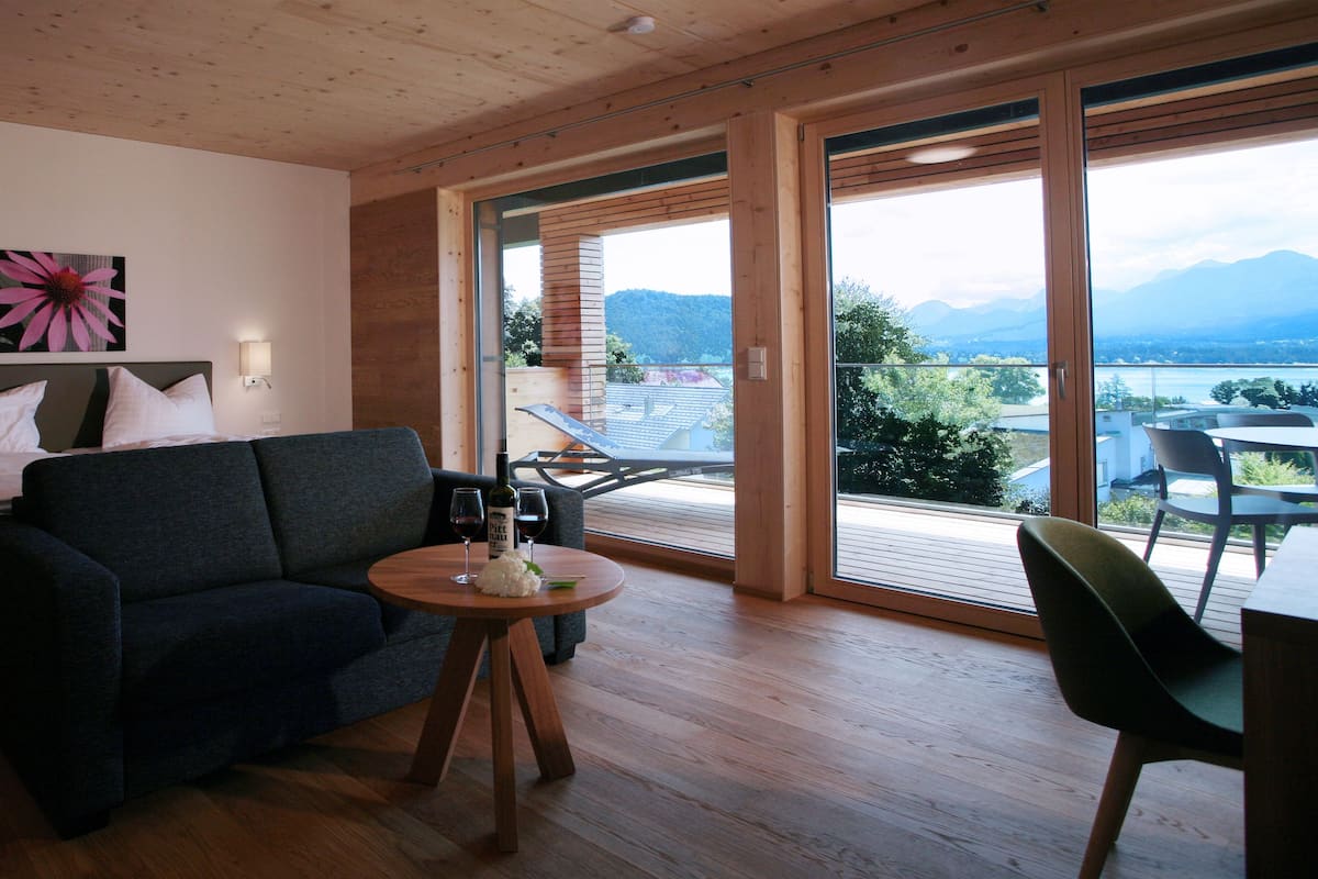 Quarto moderno com sofá, mesa redonda pequena e uma garrafa de vinho, e vista para um lago e montanhas através de grandes portas de vidro.