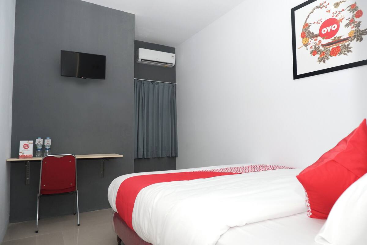 A hotel room with a bed, a desk with a chair, an air conditioner, and a flat-screen TV mounted on the wall.