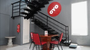 A modern interior with a staircase, a wooden table with red chairs, and a large red logo on the wall. - OYO 682 Ceria Homestay Syariah (Semarang)