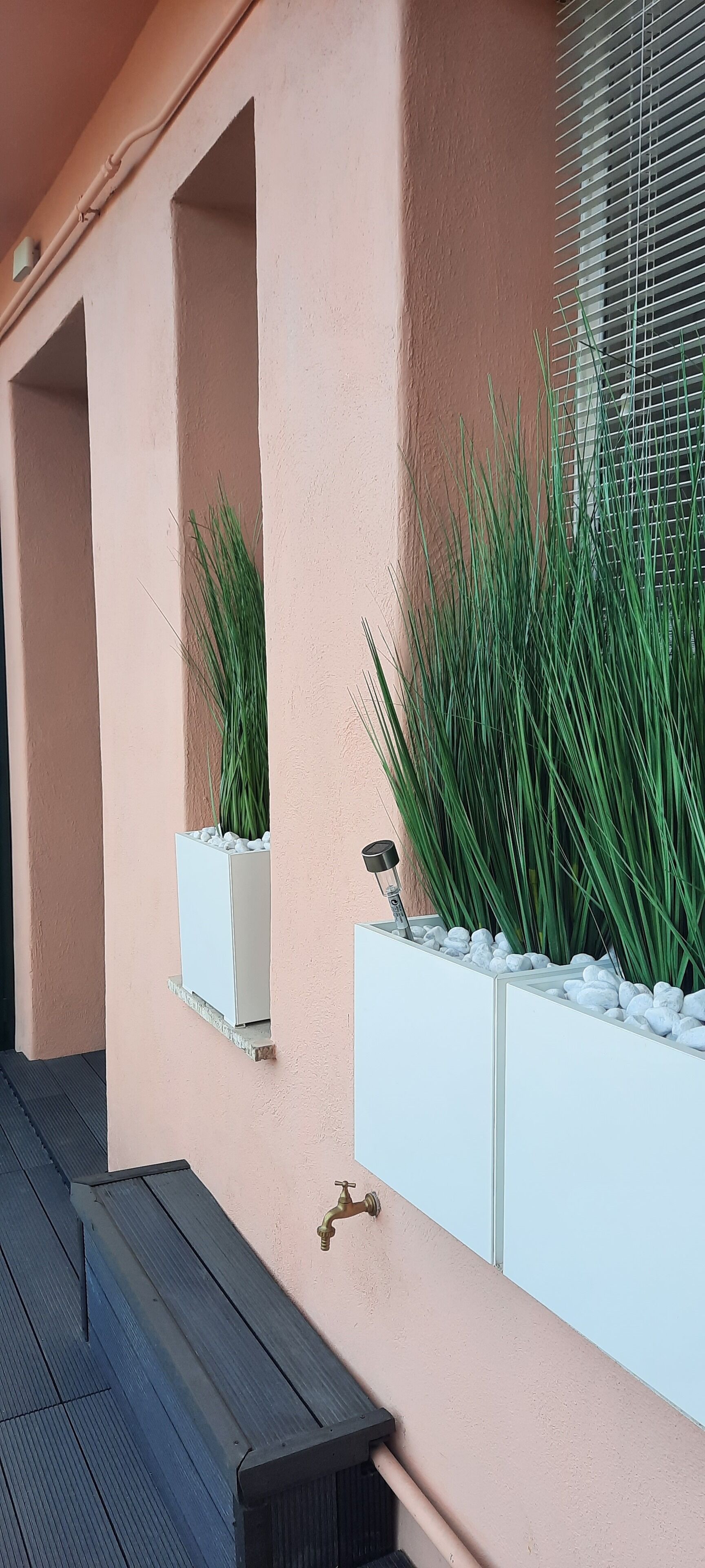 A wall with two rectangular planters containing green plants and white pebbles, a metal railing, and a black bench.