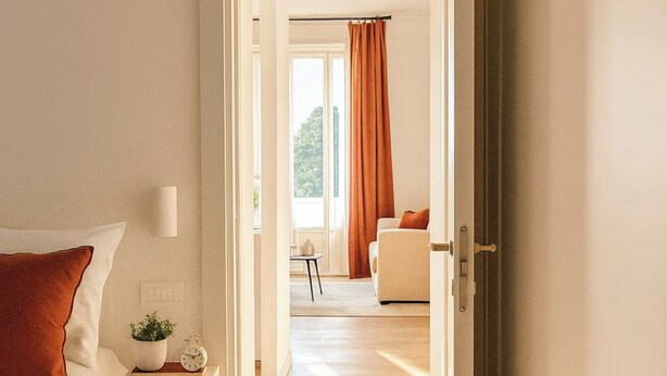 A modern bedroom with a bed, bedside table, and a bench. A hallway leads to a living area with a sofa and a window with curtains. - Penthouse with Rome View, Balconies, Two Bathrooms & Metro (Roma)