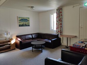 A room with a brown leather sofa, a small round table, a wooden side table, a painting on the wall, a window with curtains, and a door with an exit sign. - Sólbrekka Guesthouse (Fjardabyggd)