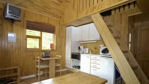 A wooden cabin interior with a kitchen, a dining table, a TV mounted on the wall, and a staircase.