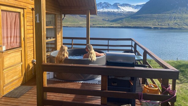 A wooden cabin with a hot tub on a deck overlooking a lake and mountains.