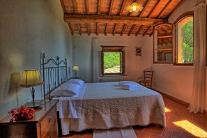 Une chambre avec un plafond en bois, un lit recouvert de draps blancs, une table de chevet avec une lampe, une fenêtre donnant sur la verdure et une chaise.
