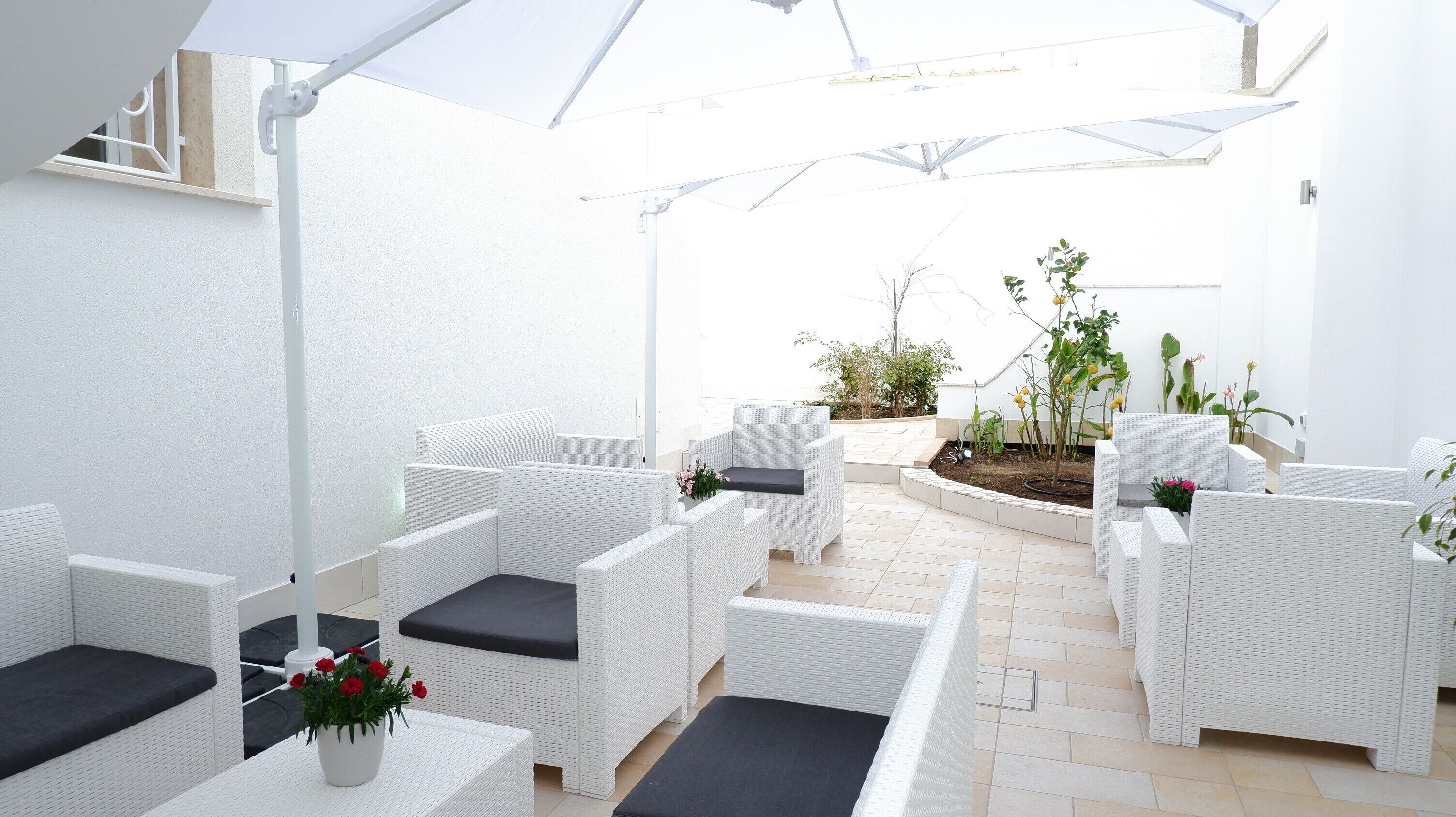 Un espace patio avec des meubles en osier blancs, un petit jardin et un mur blanc.