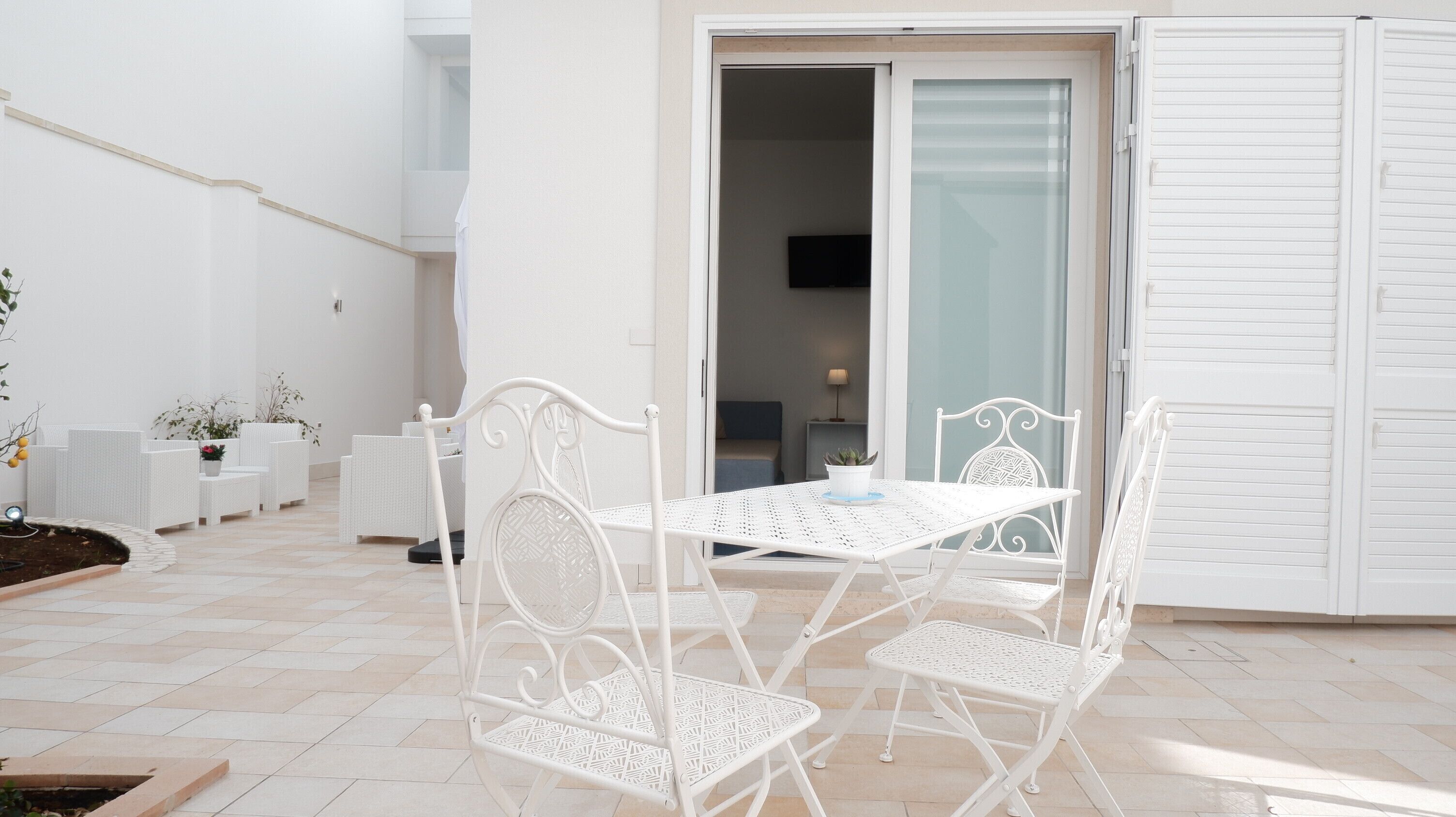 A white outdoor patio with a table and chairs, a small garden area, and a sliding glass door leading to an interior room.