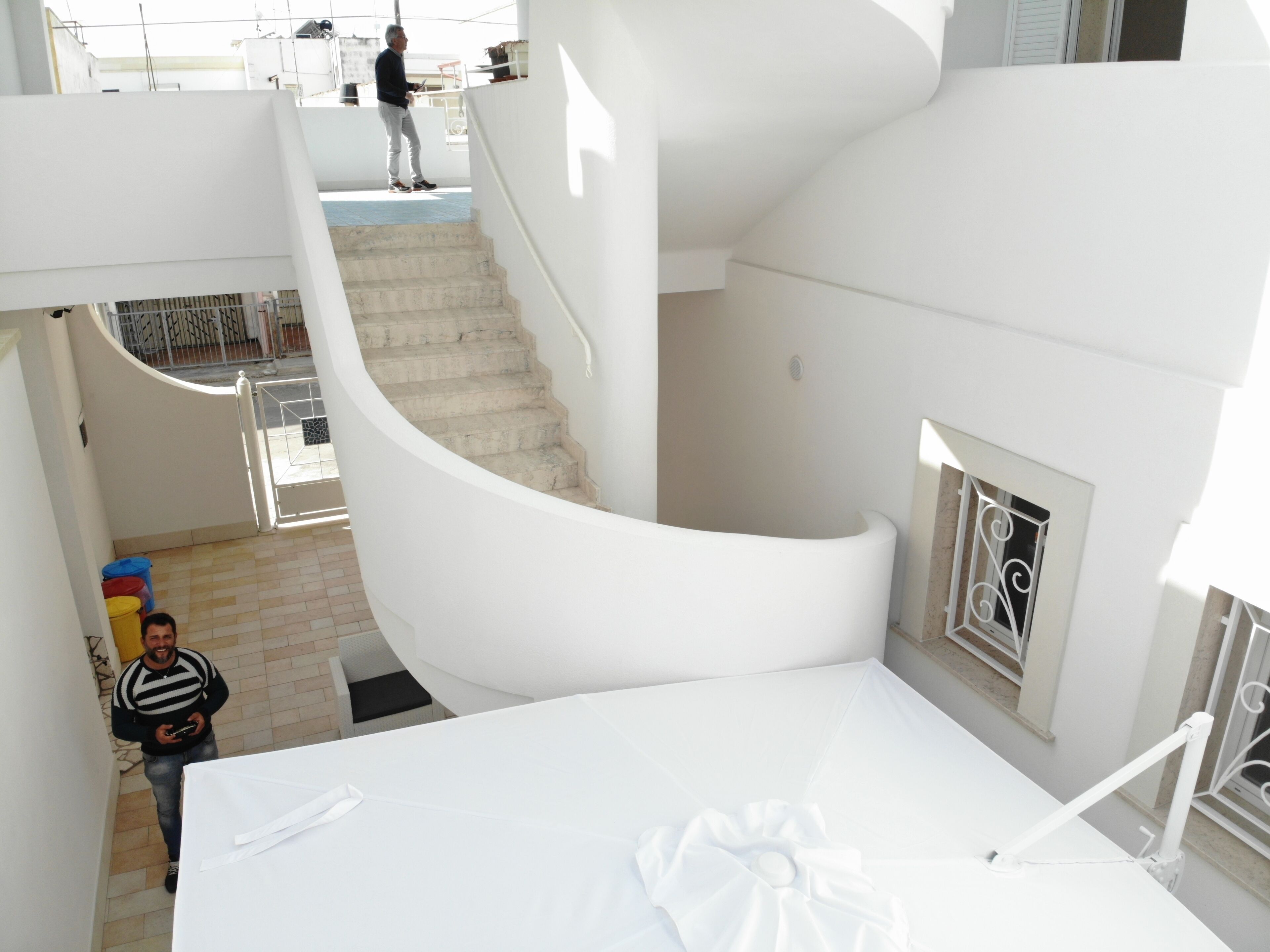 Un interno bianco con una scala a forma di ferro di cavallo, vista su un balcone, e una persona che tiene in mano un telefono.