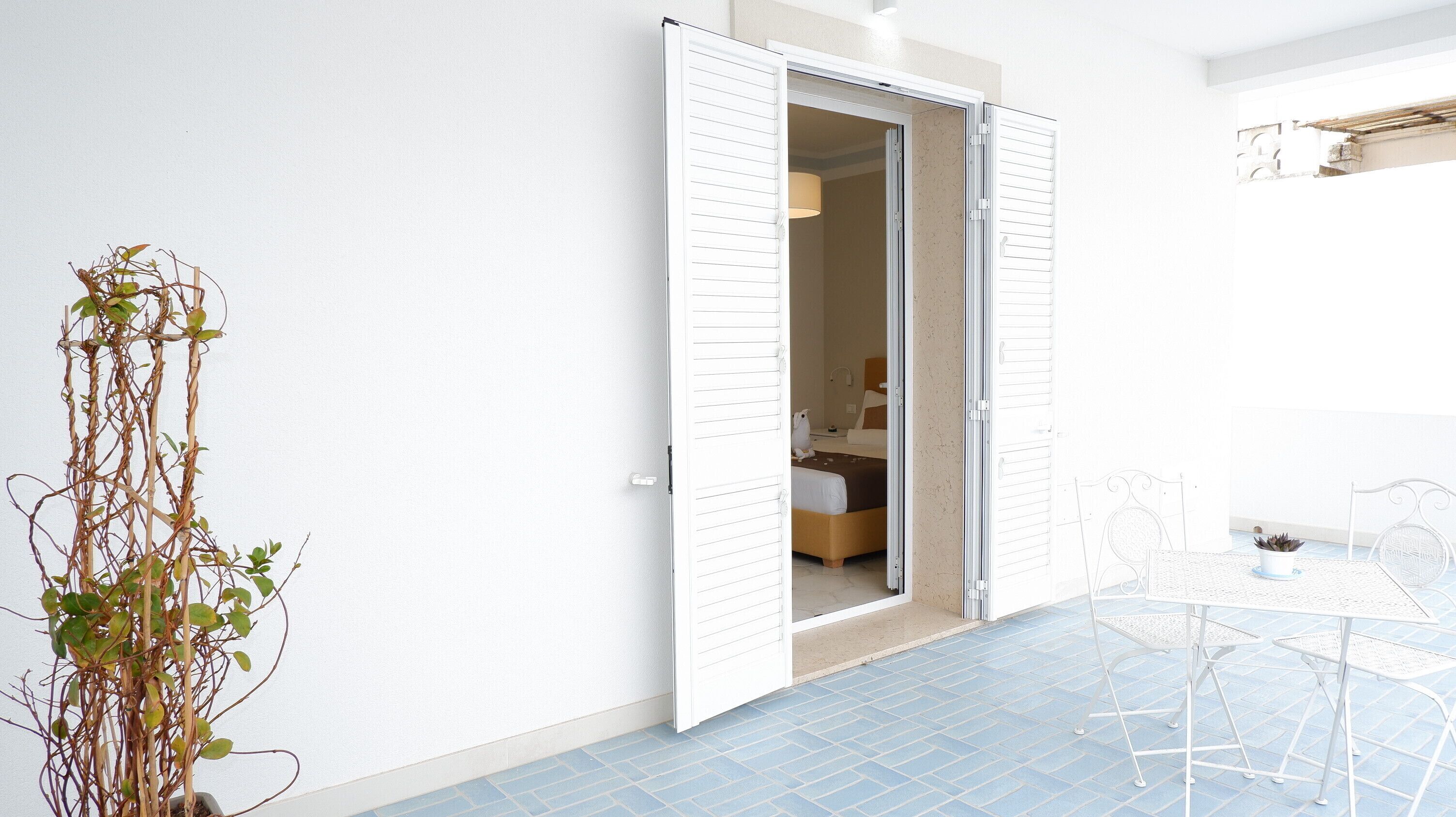 Ein Zimmer mit einem Bett, einer weißen Tür, einer Zimmerpflanze und einem kleinen Tisch mit Stühlen.