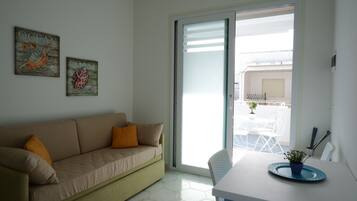 Sala de estar con un sofá beige, dos cojines, una mesa de comedor con un mantel individual azul y una planta en maceta, y un ventanal corredizo que da a un balcón con mobiliario exterior.