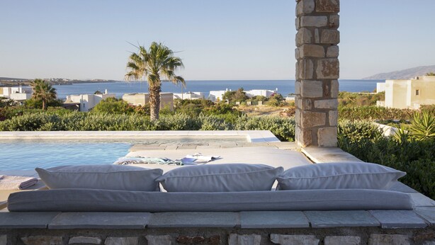 Área ao lado da piscina com um pilar de pedra, cama com almofadas e vista para o mar e edifícios ao longe.