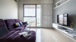 Una sala de estar moderna con un sofá morado, un televisor de pantalla plana sobre un soporte blanco y un ventanal con vistas a la ciudad.