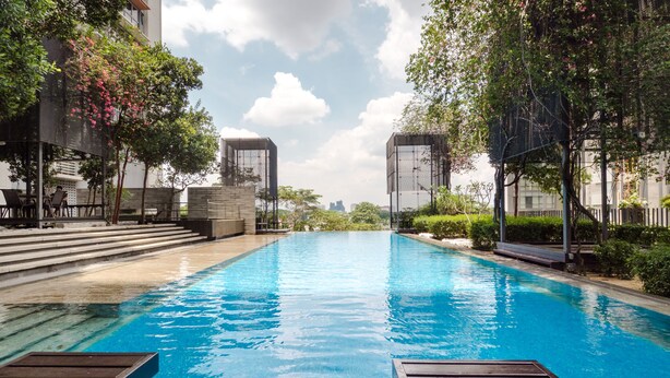 Piscina con sillones de madera, rodeada de vegetación y edificios modernos.