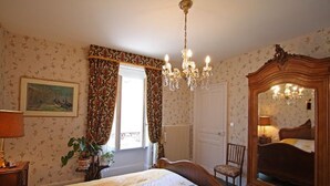 Un dormitorio con cama, cómoda, silla, candelabro y ventana con cortinas.