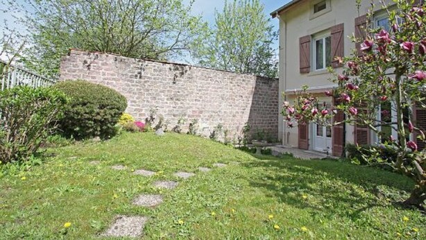 Un muro de piedra, un césped con piedras para pisar y un edificio con contraventanas y chimenea.