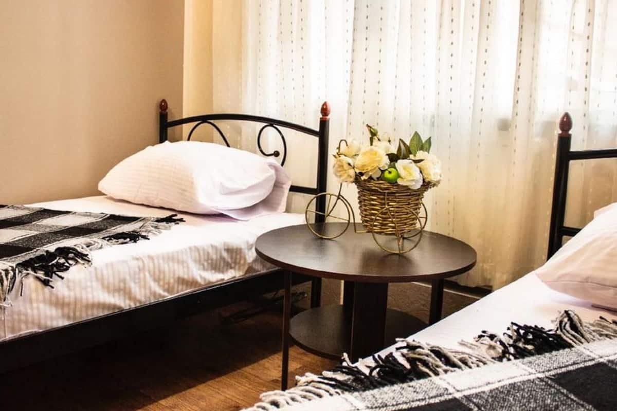 A bedroom with two single beds, a small round table with a basket of flowers, and a window with sheer curtains.
