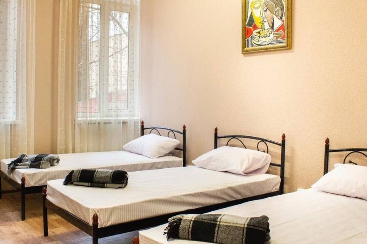 A room with three single beds, each with a white mattress and a black headboard. There is a window with sheer curtains, and a framed artwork is visible on the wall.