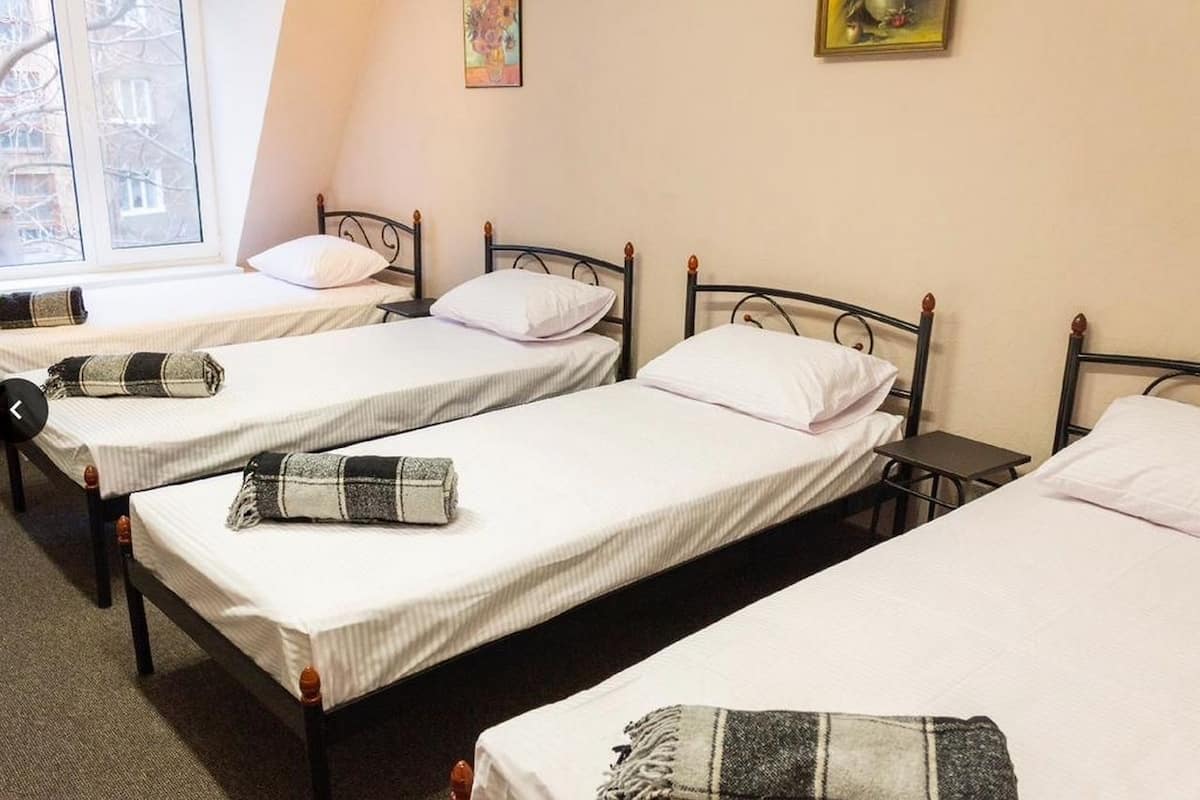 A dormitory room with three single beds, each with a white mattress and a black metal frame. There is a small table between the beds, and a window on the left side of the room.