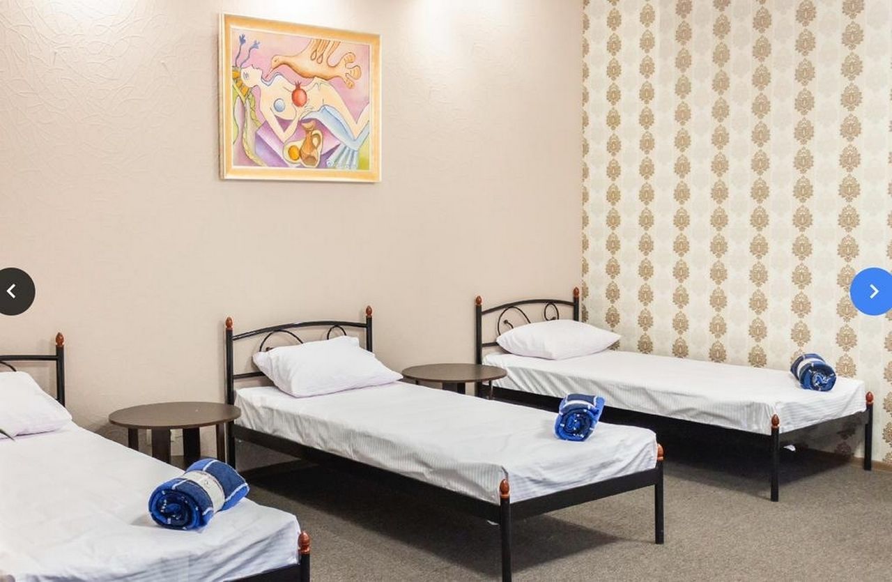 Una habitación con tres camas individuales, cada una con una almohada y una toalla enrollada. Hay una mesita redonda pequeña entre las camas. Un cuadro llamativo adorna la pared sobre las camas.