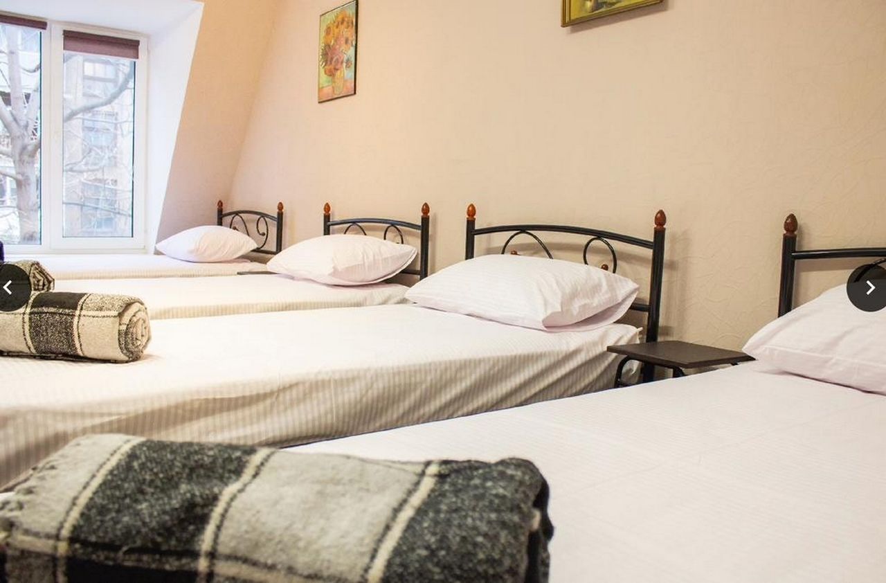 A hotel room with three single beds, a window with a view of buildings, and a painting on the wall.