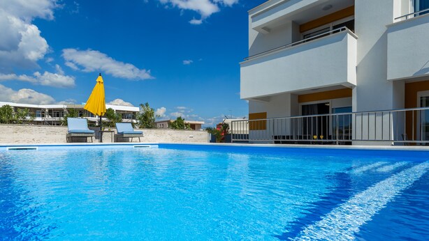 Piscina com espreguiçadeiras e um guarda-sol amarelo, cercada por um edifício branco com varandas.