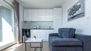 Uma sala de estar moderna com um sofá cinza, uma mesa de centro pequena e uma cozinha com armários e eletrodomésticos brancos.