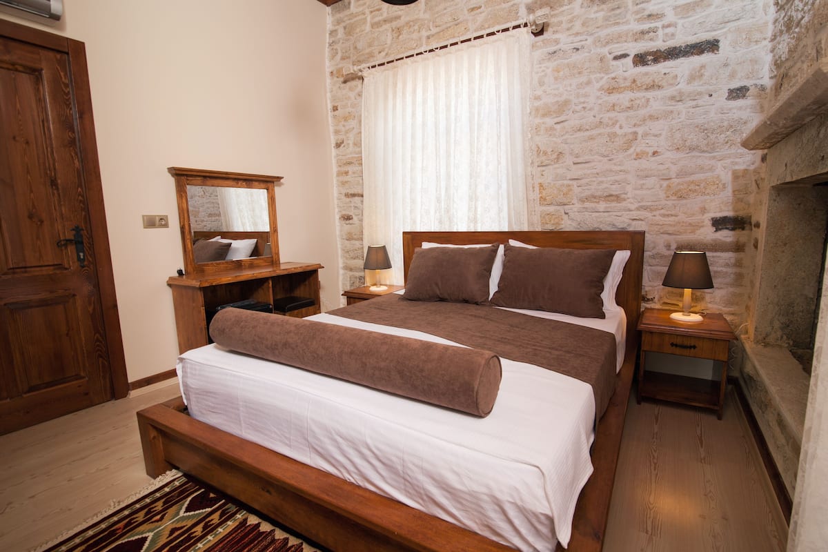 Quarto com cama de madeira, mesa de cabeceira com abajur, espelho e parede de pedra.