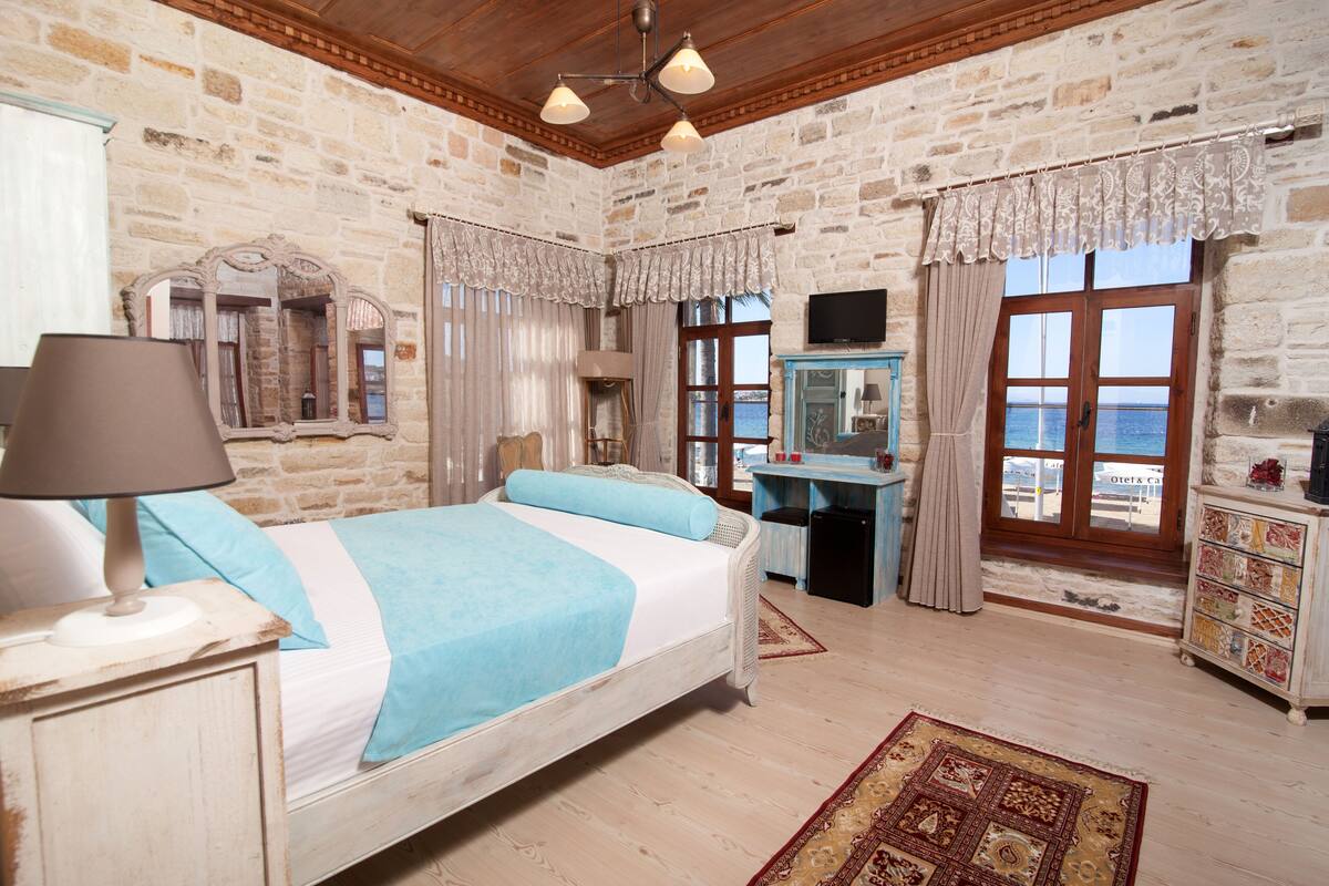 Quarto com cama, mesa de cabeceira, luminária, cômoda e vista para o mar pela janela.