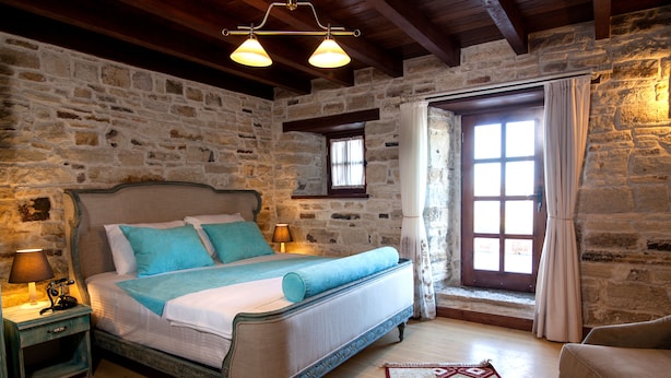 A bedroom with a stone wall, a bed with blue bedding, a nightstand with a lamp, and a window with curtains. - 1922 Hotel (Foça)