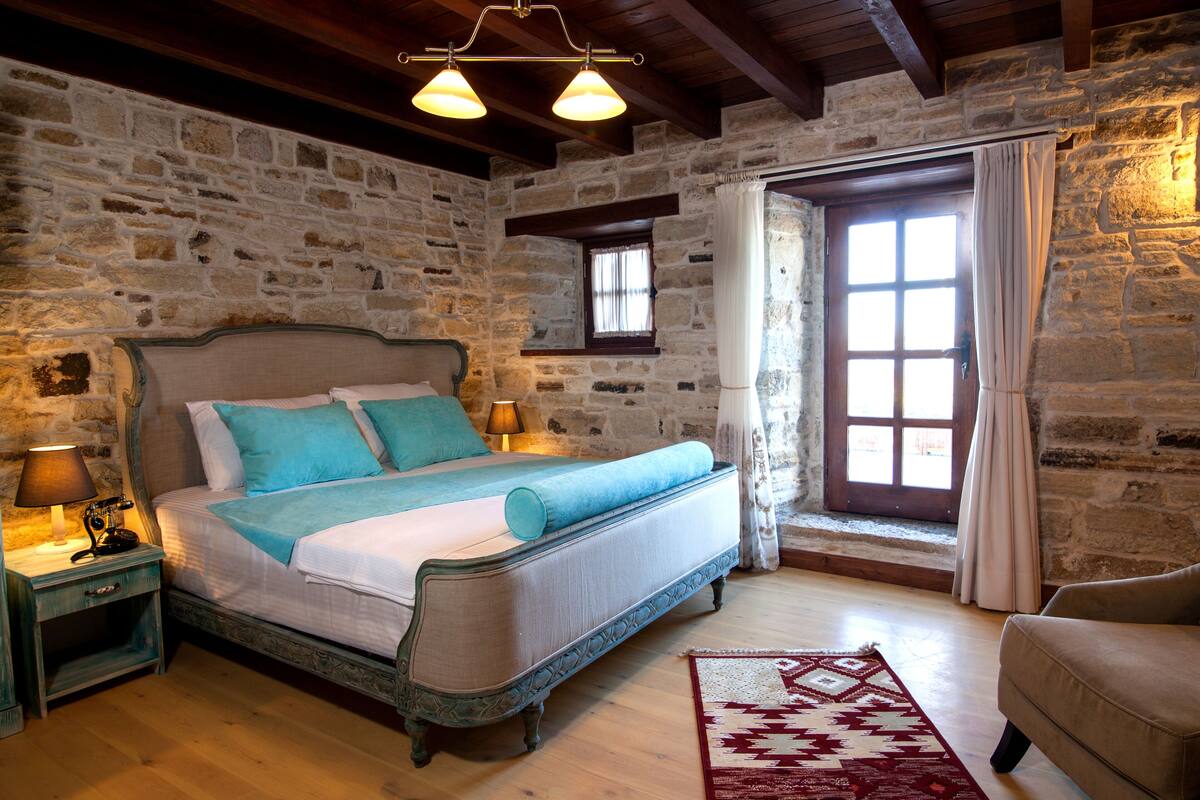 Quarto com parede de pedra, cama com roupa de cama azul, mesa de cabeceira com abajur e janela com cortinas.