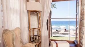 A room with a large window offering a view of the beach, a vintage armchair, a small table with a lamp, and a patterned rug. - 1922 Hotel (Foça)
