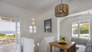 A modern room with a wooden table, a hanging light fixture, a framed picture, and a view of the sea through an open door.