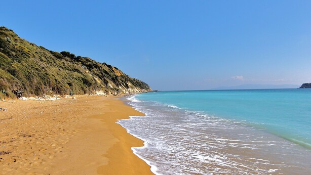 Ein Strand mit klarem, blauem Wasser, sandigem Ufer und einer hügeligen Landschaft im Hintergrund.