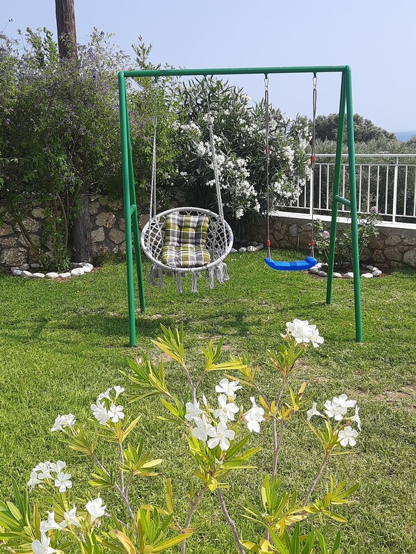 Une balançoire avec un siège en osier et une balançoire bleue, entourée d’un mur de pierre et de plantes en fleurs.