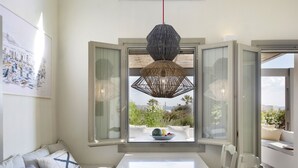 Un coin repas avec une table blanche, une fenêtre ouverte donnant sur un paysage verdoyant et un luminaire suspendu en fibres tressées.