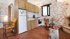 A compact kitchen with wooden cabinets, a white refrigerator, a stove, and a dining area with a wooden chair and a table. - Anemos Studio (Zakynthos)