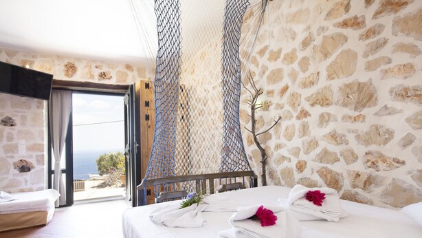 A bedroom with a stone wall, a bed with white linens, and a view of the sea through a glass door. - Anemos Studio (Zakynthos)