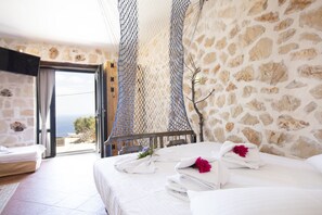 A bedroom with a stone wall, a bed with white linens, and a view of the sea through a glass door.