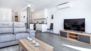 Un salon moderne avec un canapé gris, une table basse en bois sur laquelle sont posées des tasses, un téléviseur sur un meuble en bois, et une cuisine en arrière-plan.