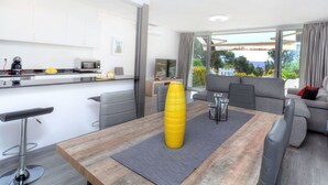 Un salon moderne avec un canapé, une table à manger agrémentée d’un vase jaune, et un coin cuisine doté de meubles blancs et d’une machine à café.
