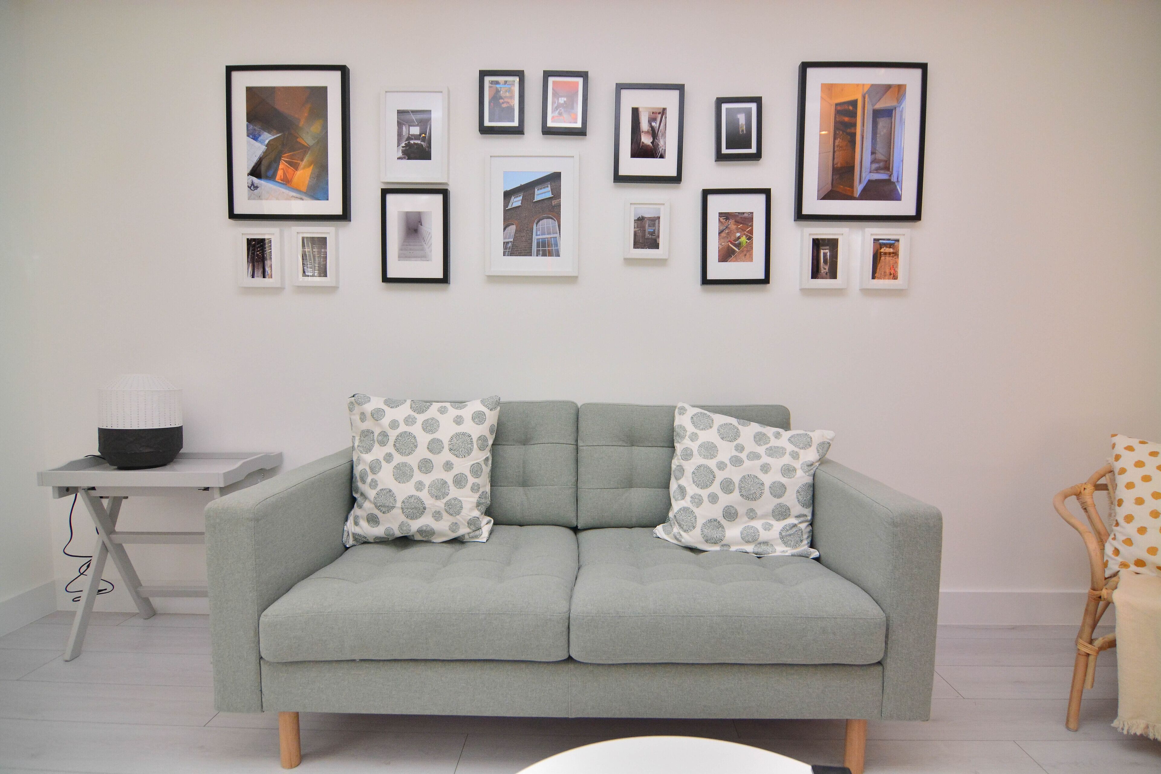 A living room with a grey sofa, two patterned pillows, a small table with a lamp, and a framed picture collection on the wall.