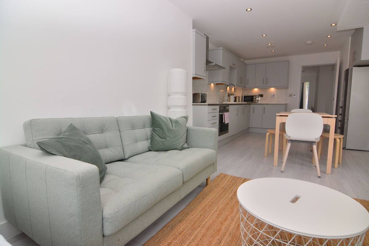 A modern living room with a light grey sofa, a small round table, and a kitchen area with white cabinets and a dining table.