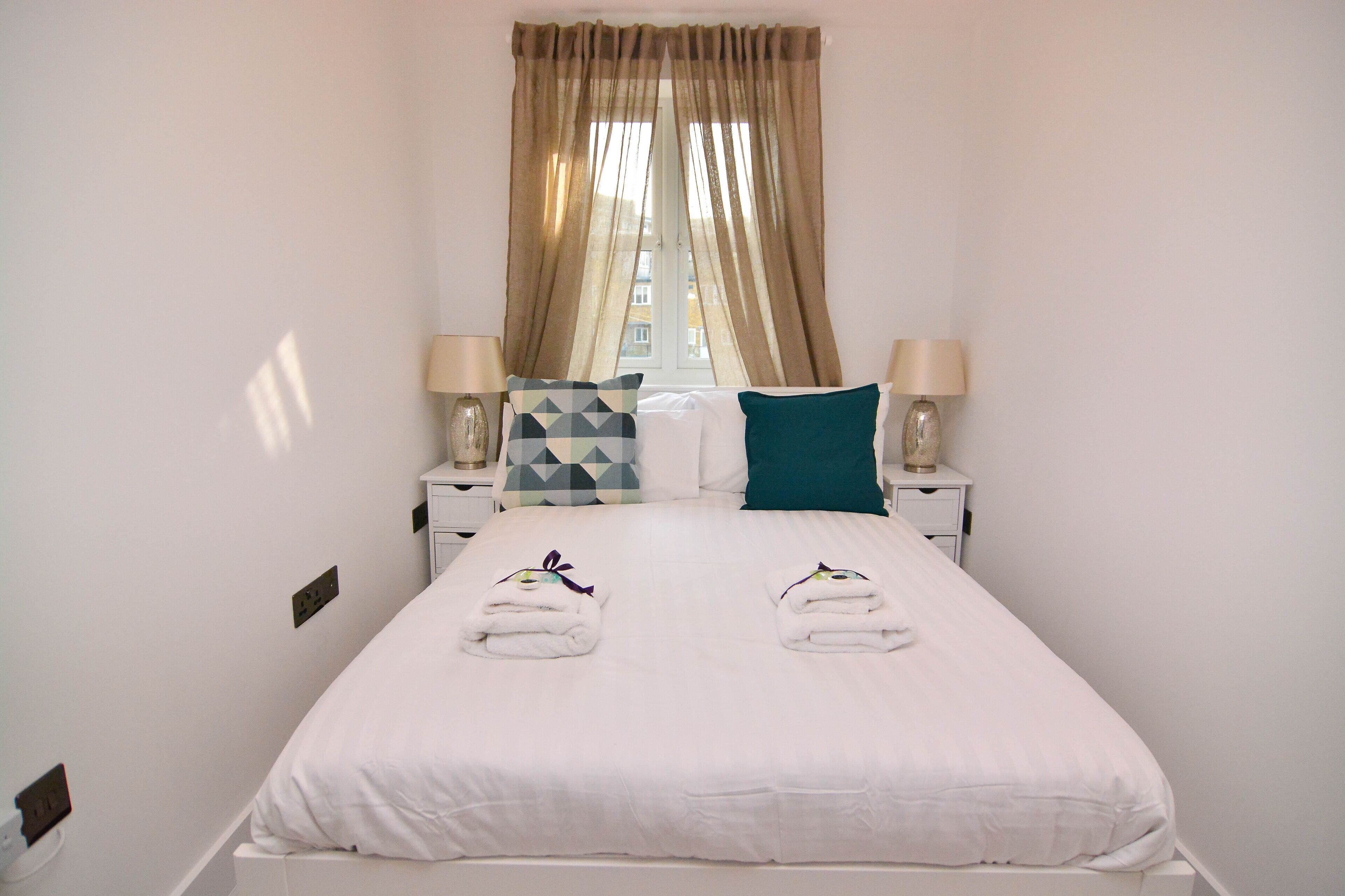 A neatly made bed with white linens, two pillows, and two folded towels. There are two bedside tables with lamps and a window with curtains.